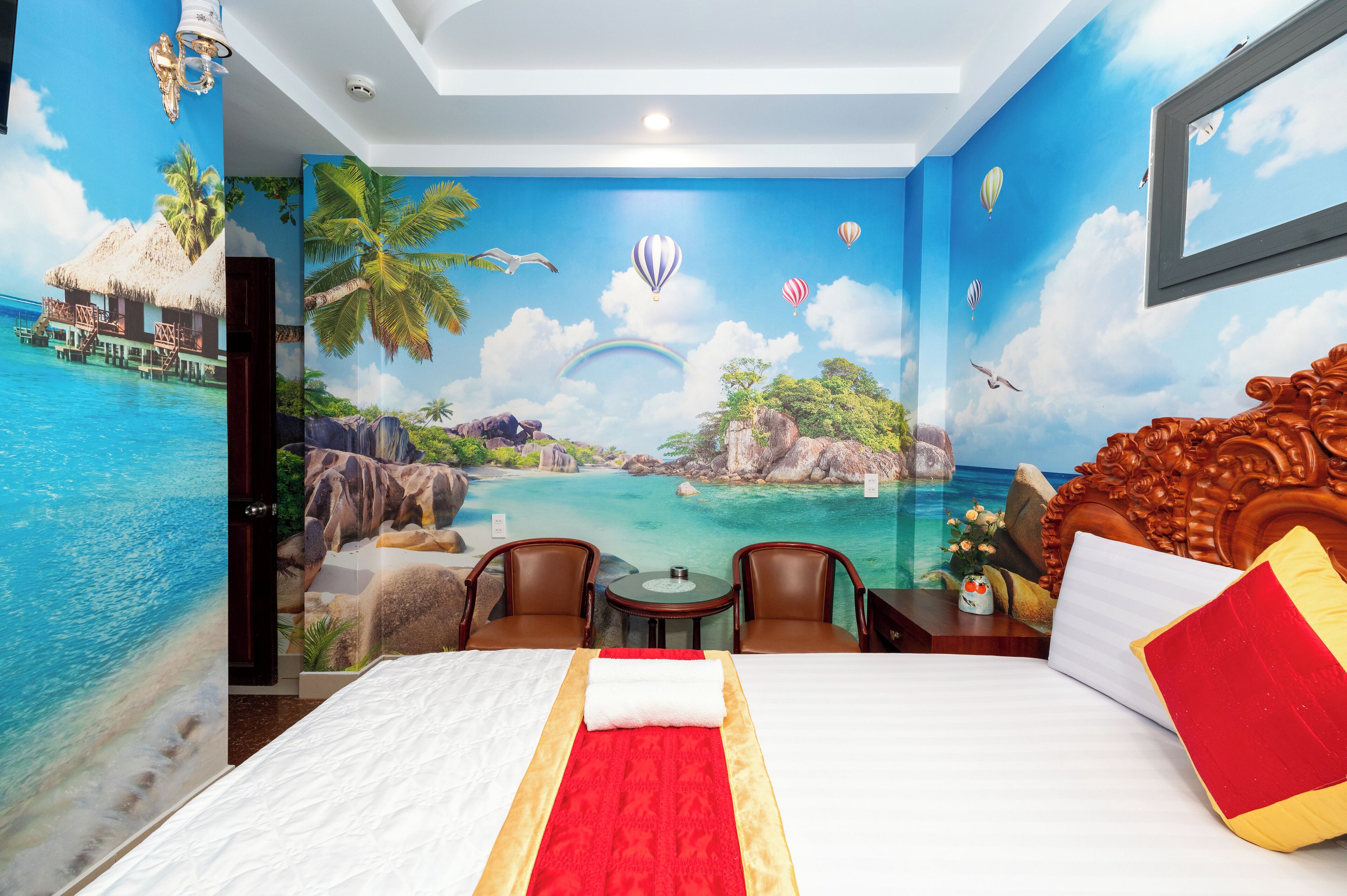 Hotelkamer met een muurschildering van een tropisch strand, een bed met een rood-witte plaid, twee stoelen en een kleine tafel.
