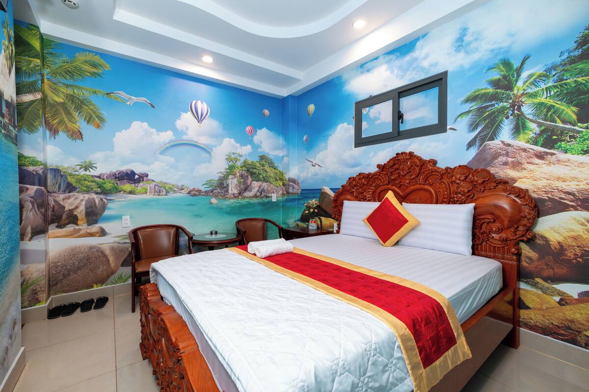 A bedroom with a mural of a tropical beach, a wooden bed with a red and white bedspread, a chair, and a small table.