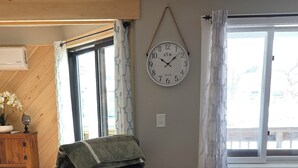 Una sala de estar con sillón de cuero y reposapiés, suelo de madera, un reloj de pared y una ventana con cortinas.