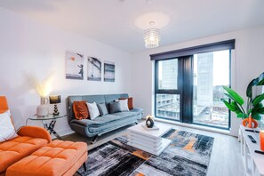 A modern living room with a sofa, coffee table, and orange armchairs. There is a large window with a view of the city. - 2BDR | Free Parking | Stylish Liverpool Apartment (Liverpool)