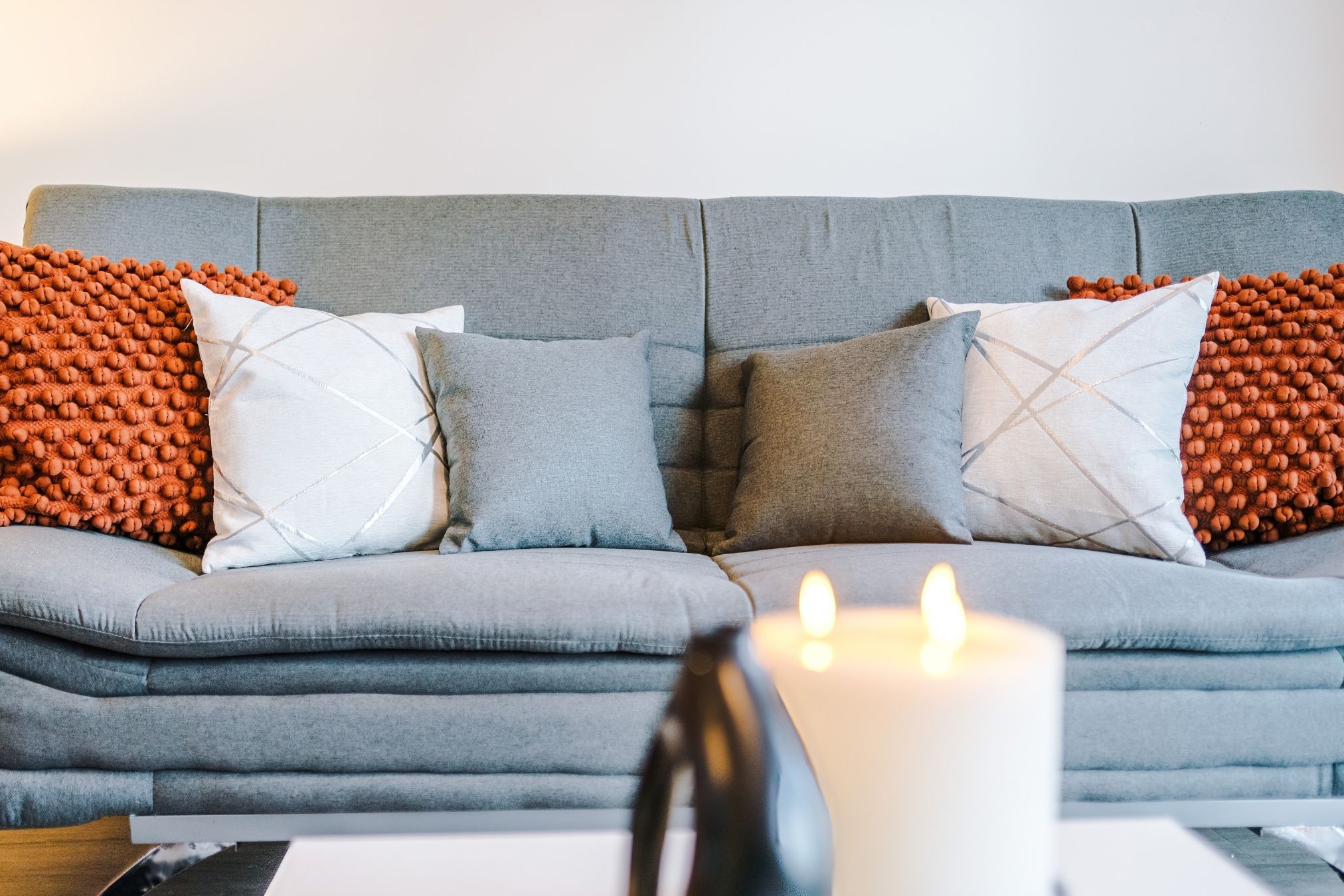 A grey sofa with multiple cushions and a white candle on a table in front.