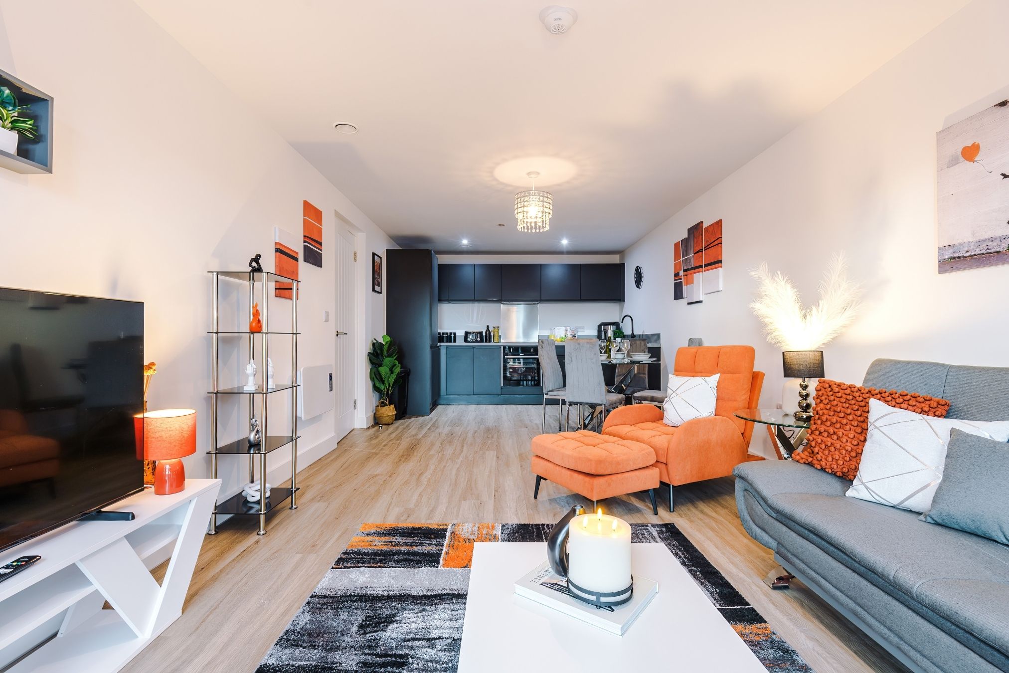 A modern living room with a sofa, armchairs, a coffee table, and a TV. There is a kitchen in the background.