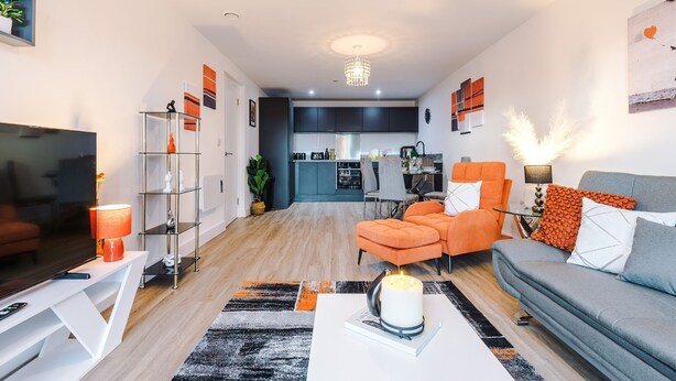 A modern living room with a sofa, armchairs, a coffee table, and a TV. There is a kitchen in the background. - 2BDR | Free Parking | Stylish Liverpool Apartment (Liverpool)