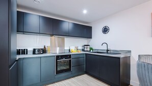 A modern kitchen with dark cabinetry, a built-in oven, a stainless steel sink, and a clock on the wall. - 2BDR | Free Parking | Stylish Liverpool Apartment (Liverpool)