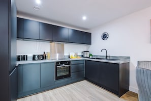 A modern kitchen with dark cabinetry, a built-in oven, a stainless steel sink, and a clock on the wall. - 2BDR | Free Parking | Stylish Liverpool Apartment (Liverpool)