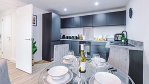 A modern kitchen with dark cabinets, a dining area with a glass table set for four, and a clock on the wall. - 2BDR | Free Parking | Stylish Liverpool Apartment (Liverpool)