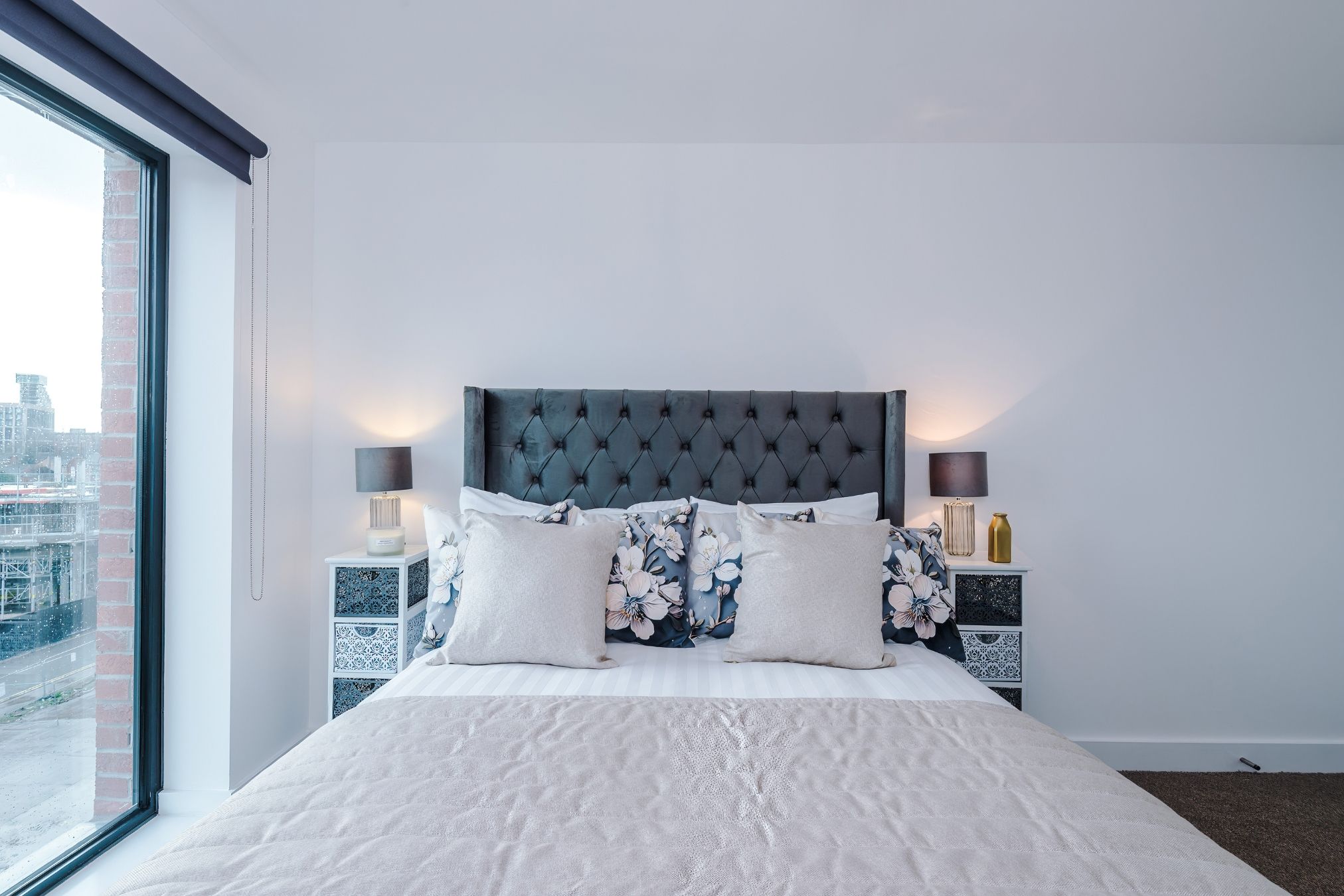 A bedroom with a large bed, two nightstands, a window with a view of buildings, and a cityscape.