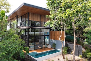 Ein modernes, zweistöckiges Haus mit Glasfassade, einer Holzterrasse und einem Swimmingpool.