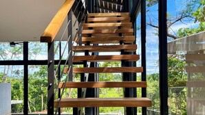 Una escalera moderna con escalones de madera y barandillas de metal que lleva a un nivel superior con amplios ventanales que ofrecen vistas a la vegetación.