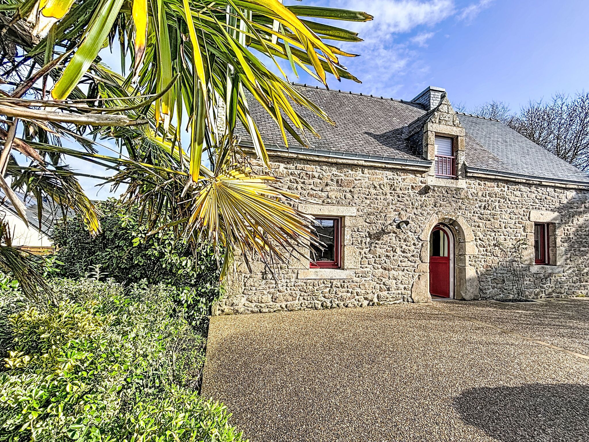 A stone building with a red door and two windows, surrounded by greenery.