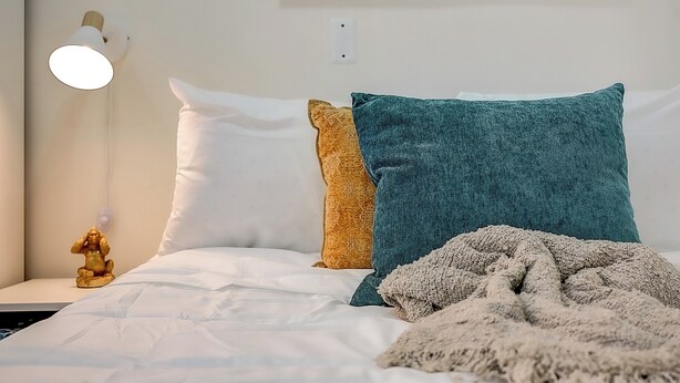 Una cama bien hecha con ropa de cama blanca, un cojín verde azulado, un cojín color mostaza y una manta color beige.