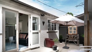 Eine Terrassenfläche mit einer Schiebetür, die zu einem Raum führt, ein rotes Stuhl, ein kleiner Topfpflanze und ein weißer Sonnenschirm, der Schatten spendet.