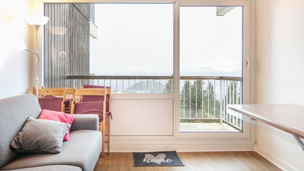 A room with a sofa, a small table, a chair, a window with a view of mountains, and a balcony with a railing. - Cozy Mountain Base Near Bachat Bouloud (Chamrousse)