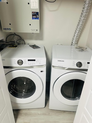 Zwei Frontlader-Waschmaschinen, eine mit dem Aufdruck 'Hero' und die andere von Samsung, mit einem Wasserzähler und einem Schlauch im Hintergrund.
