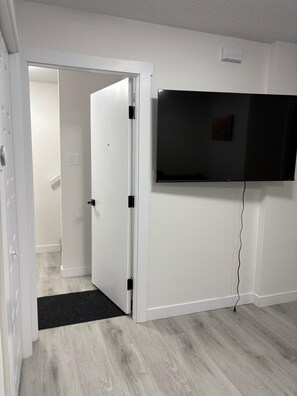 Ein Zimmer mit einer weißen Tür, einer schwarzen Matte am Eingang, einem an der Wand befestigten Fernseher und Holzdielen.