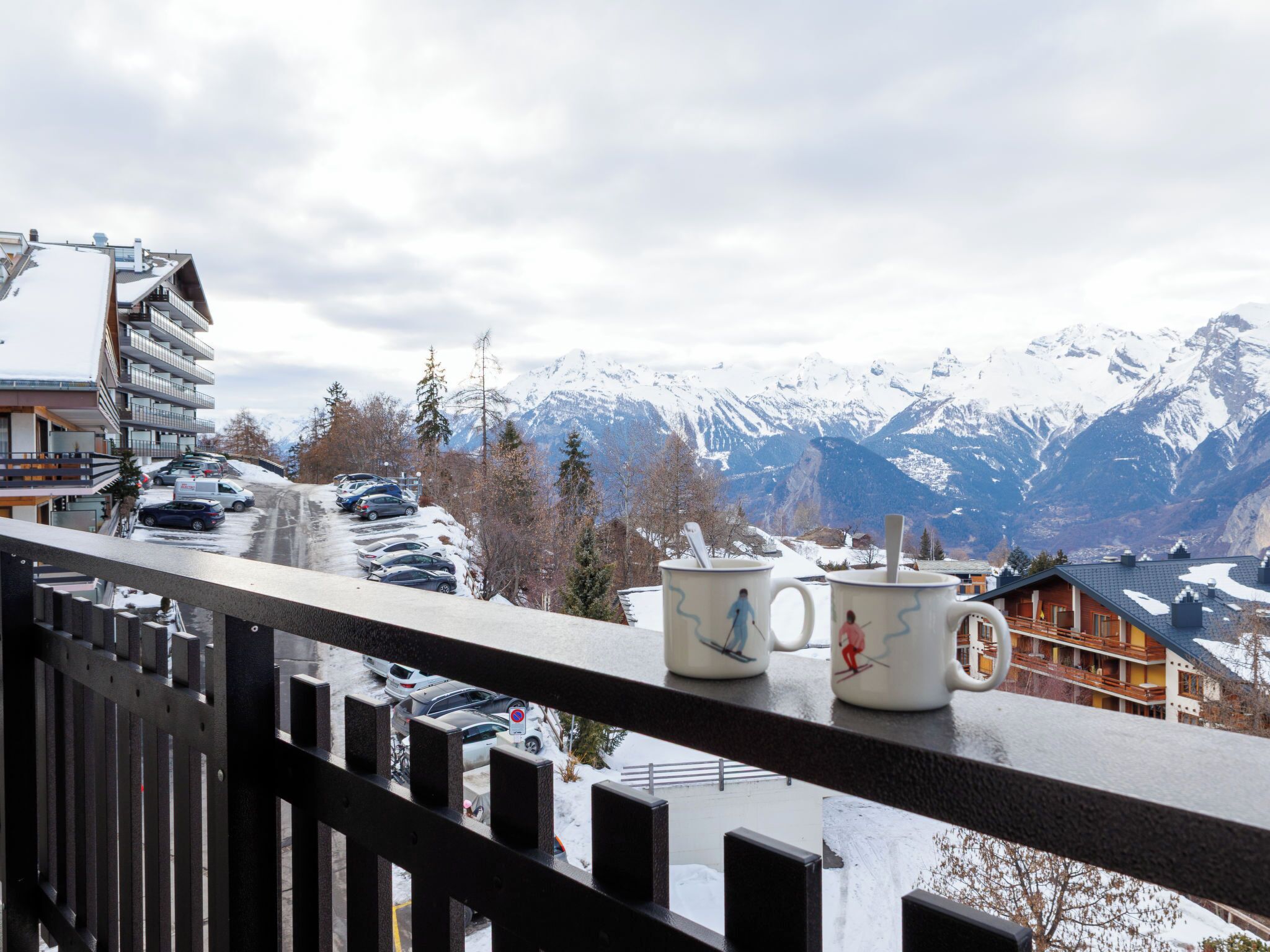 Un balcon avec deux tasses, une chaîne de montagnes enneigée et, au loin, un bâtiment doté de balcons.