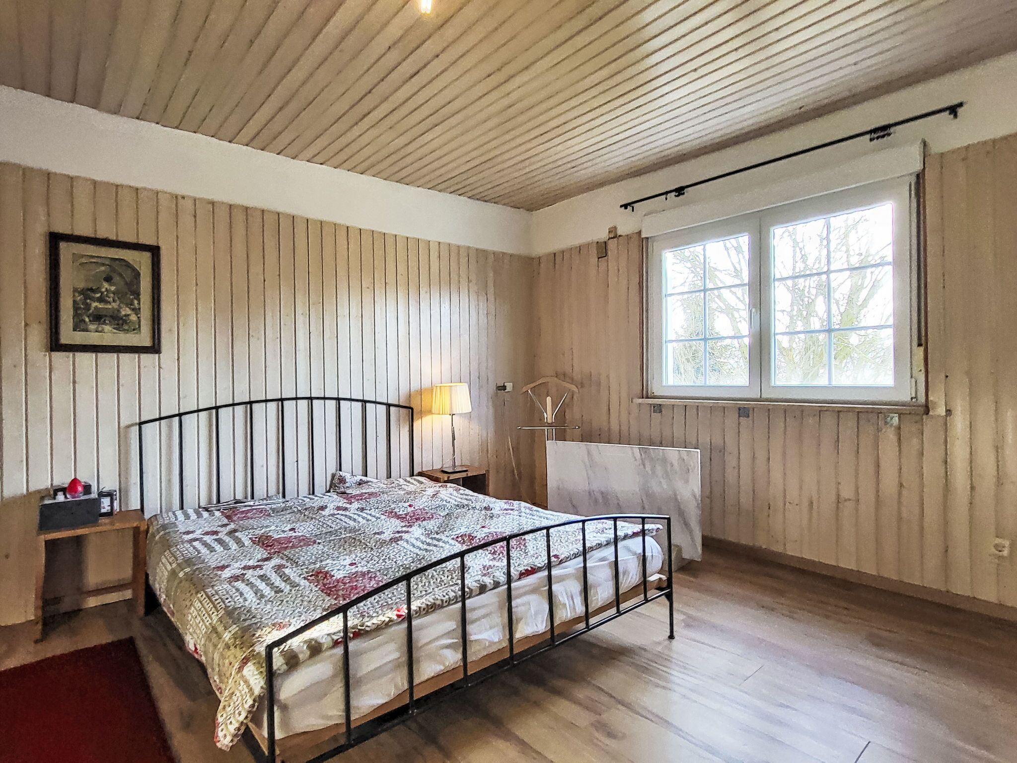 Ein Schlafzimmer mit Holzboden, einem Bett mit Metallgestell, einem Nachttisch mit Lampe, einem Fenster mit Vorhängen und einem gerahmten Bild an der Wand.