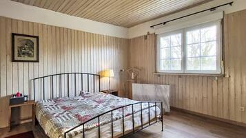 Ein Schlafzimmer mit Holzboden, einem Bett mit Metallgestell, einem Nachttisch mit Lampe, einem Fenster mit Vorhängen und einem gerahmten Bild an der Wand.