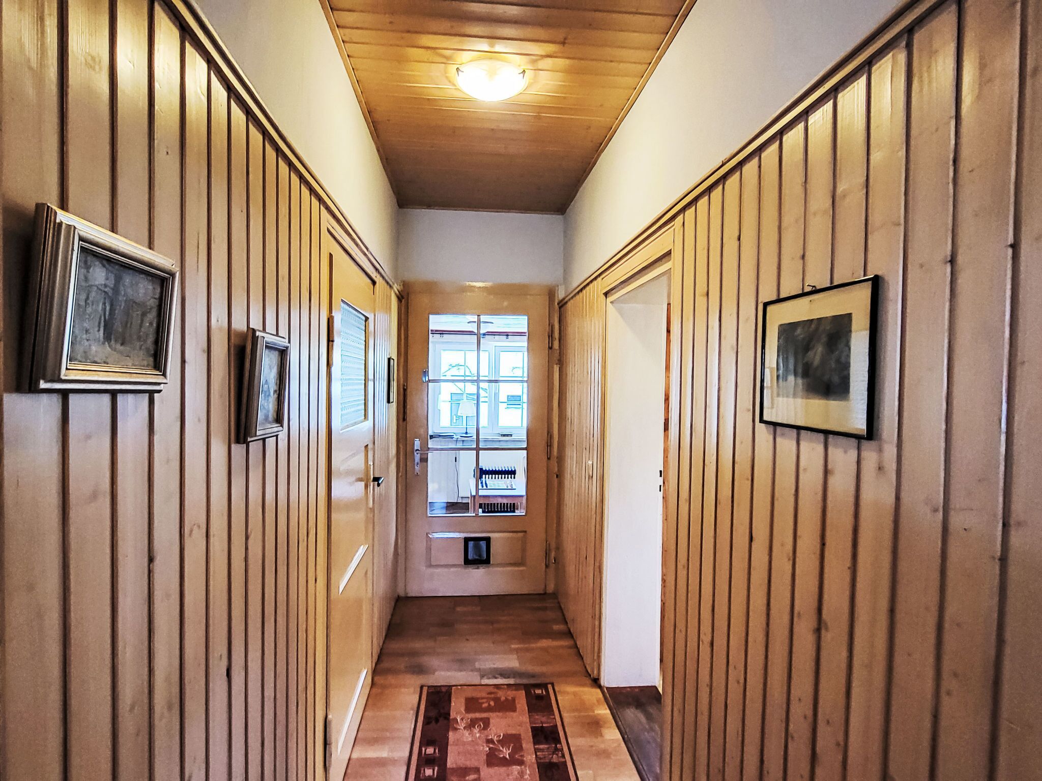Ein Korridor mit Holzwandverkleidung, gerahmten Bildern an der Wand, einem gemusterten Teppich und einem Fenster am Ende.