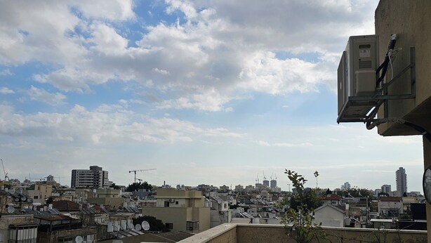 Terraza en la azotea con plantas en macetas, una antena parabólica y vistas de un paisaje urbano con edificios y aerogeneradores.