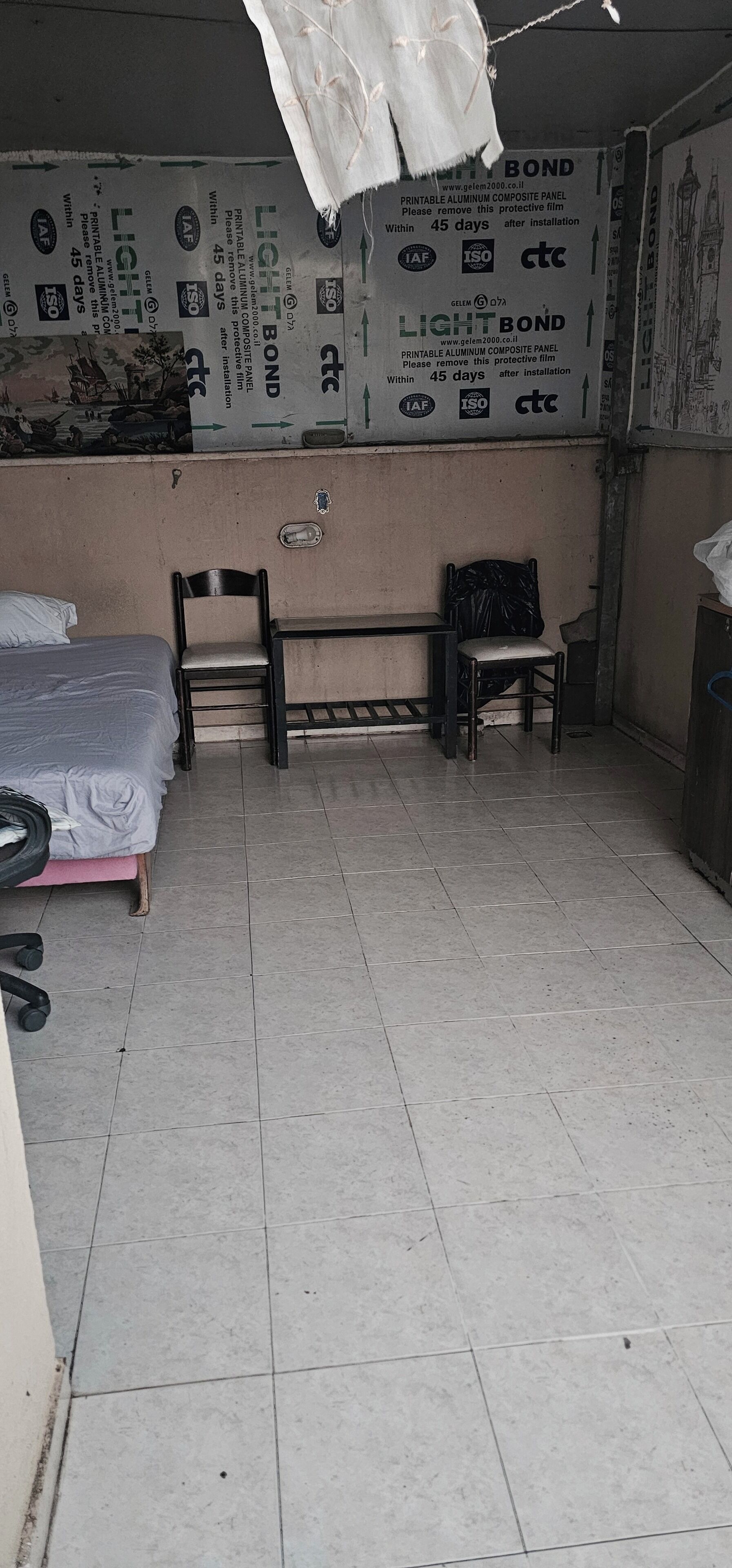 Een kleine, zwak verlichte kamer met een bed, twee stoelen en een bureau. De muren zijn bekleed met beschermfolie en voorzien van diverse logo's en tekst.