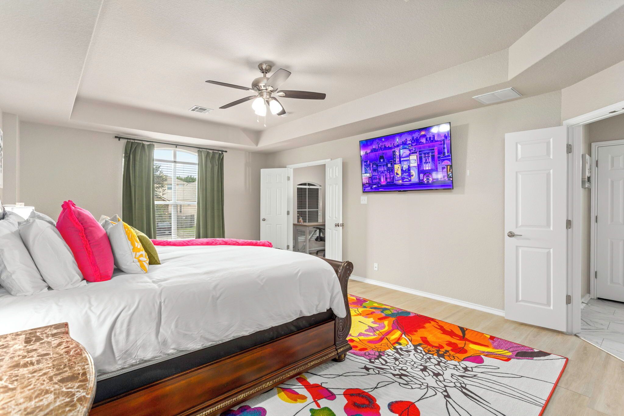 A bedroom with a bed, a ceiling fan, a TV on the wall, and a colorful rug.