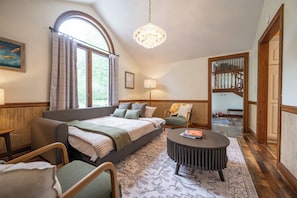 A bedroom with a large bed, a round coffee table, a sofa, and a chair. There is a window with curtains, a lamp, and a painting on the wall.