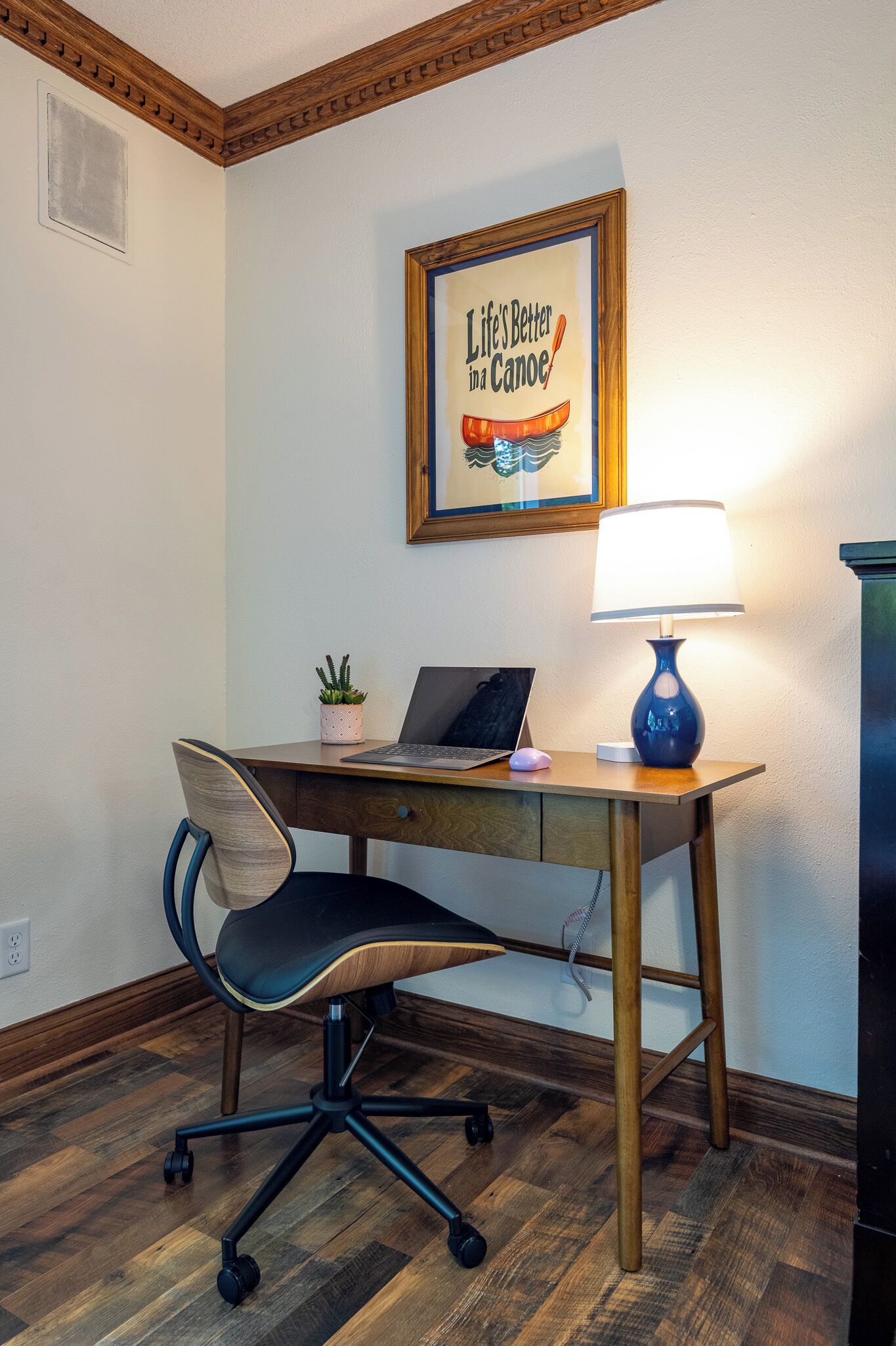 Un escritorio de madera con una laptop, una lámpara azul, una planta en maceta y una cita enmarcada en la pared.