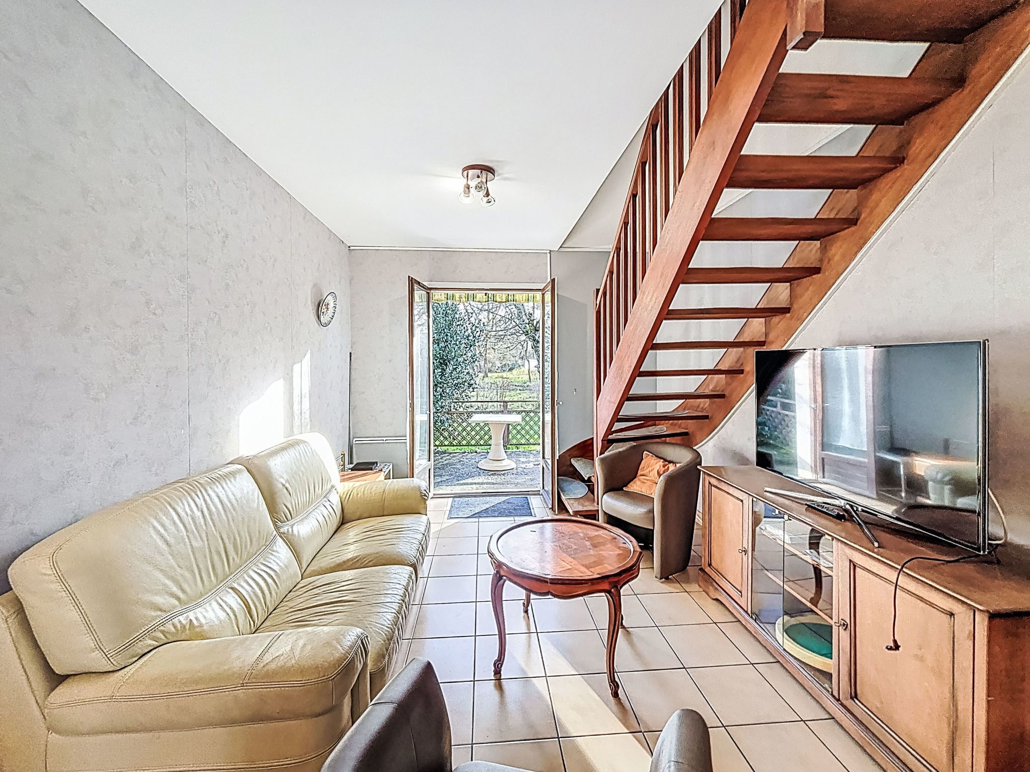 A living room with a sofa, a TV, a wooden staircase, and a view of the outdoors.
