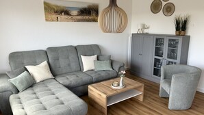 Un salon comprenant un canapé d’angle gris, une table basse en bois, un fauteuil gris, un meuble gris avec des portes en verre et une suspension.