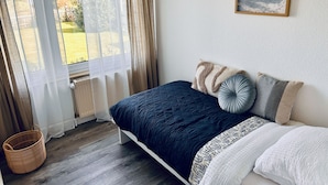 Une chambre à coucher avec un lit, une fenêtre avec des rideaux, un radiateur, un panier en osier et un tableau accroché au mur.
