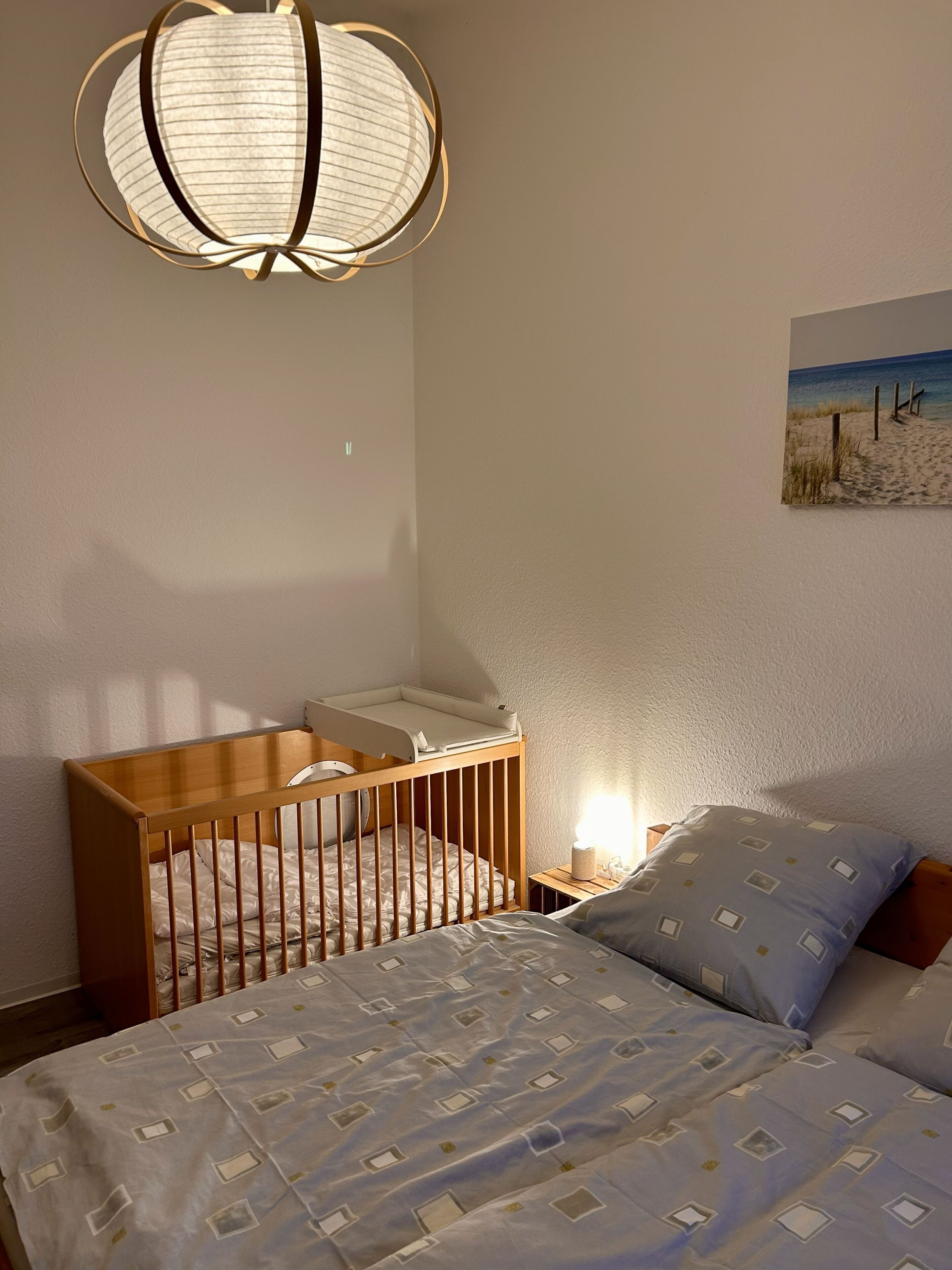 Une chambre avec un berceau, un lit avec une literie à motifs, une lampe fixée au mur et un tableau sur le thème de la plage.