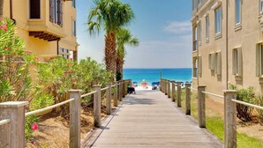 A wooden boardwalk leading to a beach with a palm tree and a building with a balcony. - SunnySide: Private beach steps away, FREE Golf Cart, Heated pool, Luxury home (Destin)