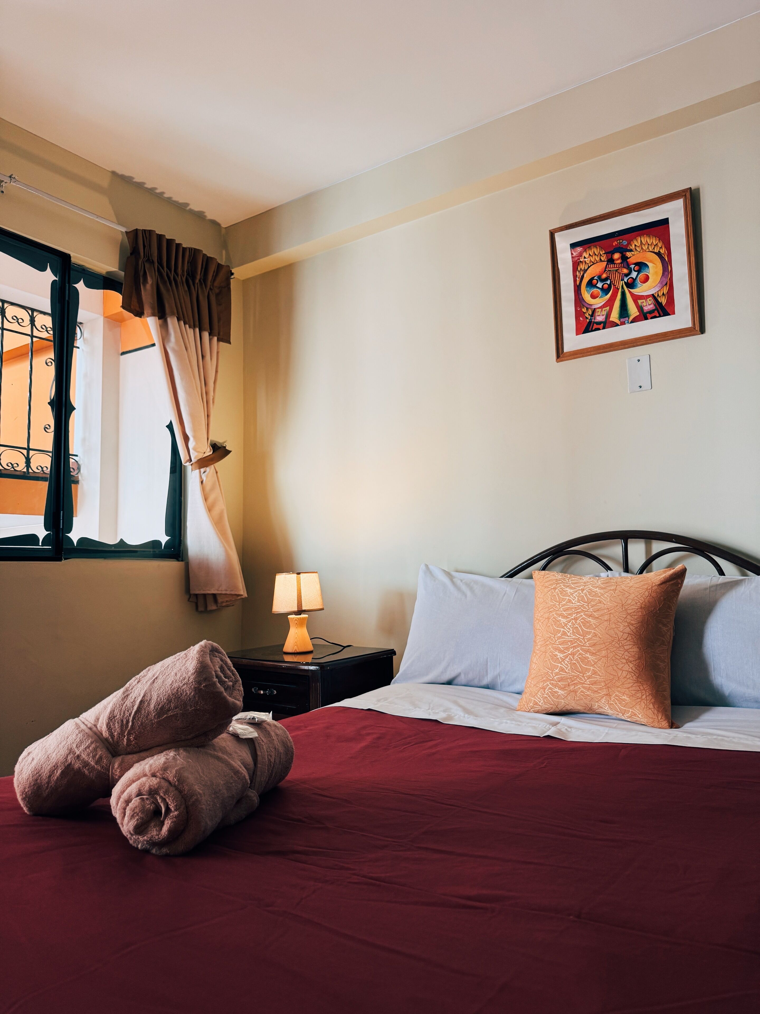 A hotel room with a neatly made bed, a bedside lamp, a window with curtains, and a wall-mounted artwork.