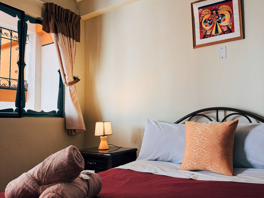 A hotel room with a neatly made bed, a bedside lamp, a window with curtains, and a wall-mounted artwork.