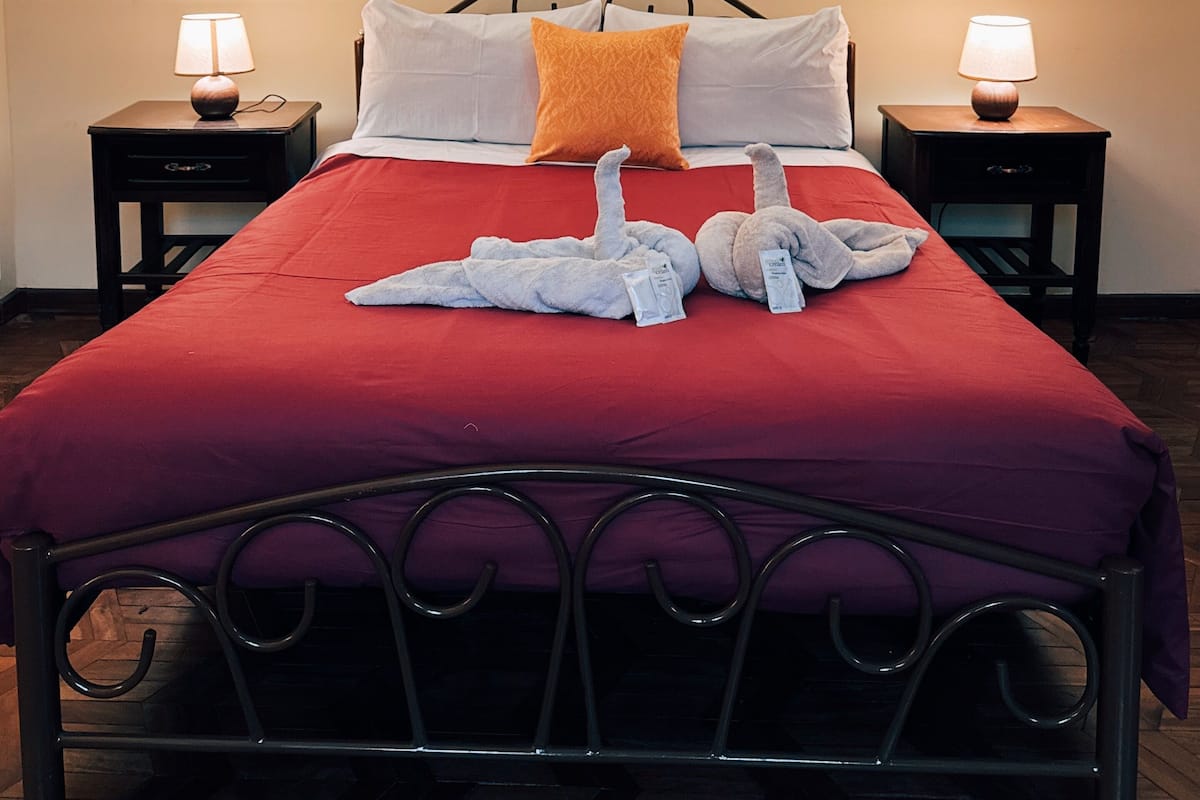 A neatly made bed with a red bedspread, two pillows, and a decorative pillow. There are two bedside tables with lamps, a framed artwork on the wall, and a wooden floor.