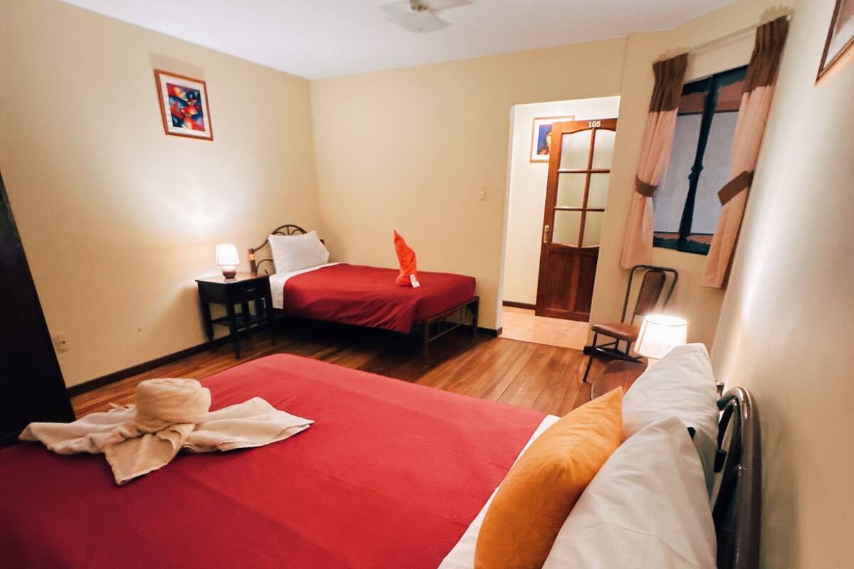 A hotel room with two beds, a small table, a nightstand, a window with curtains, and a door with a number sign.