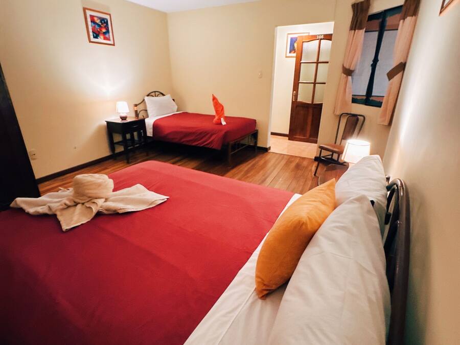 A hotel room with two beds, a small table, a nightstand, a window with curtains, and a door with a number sign.