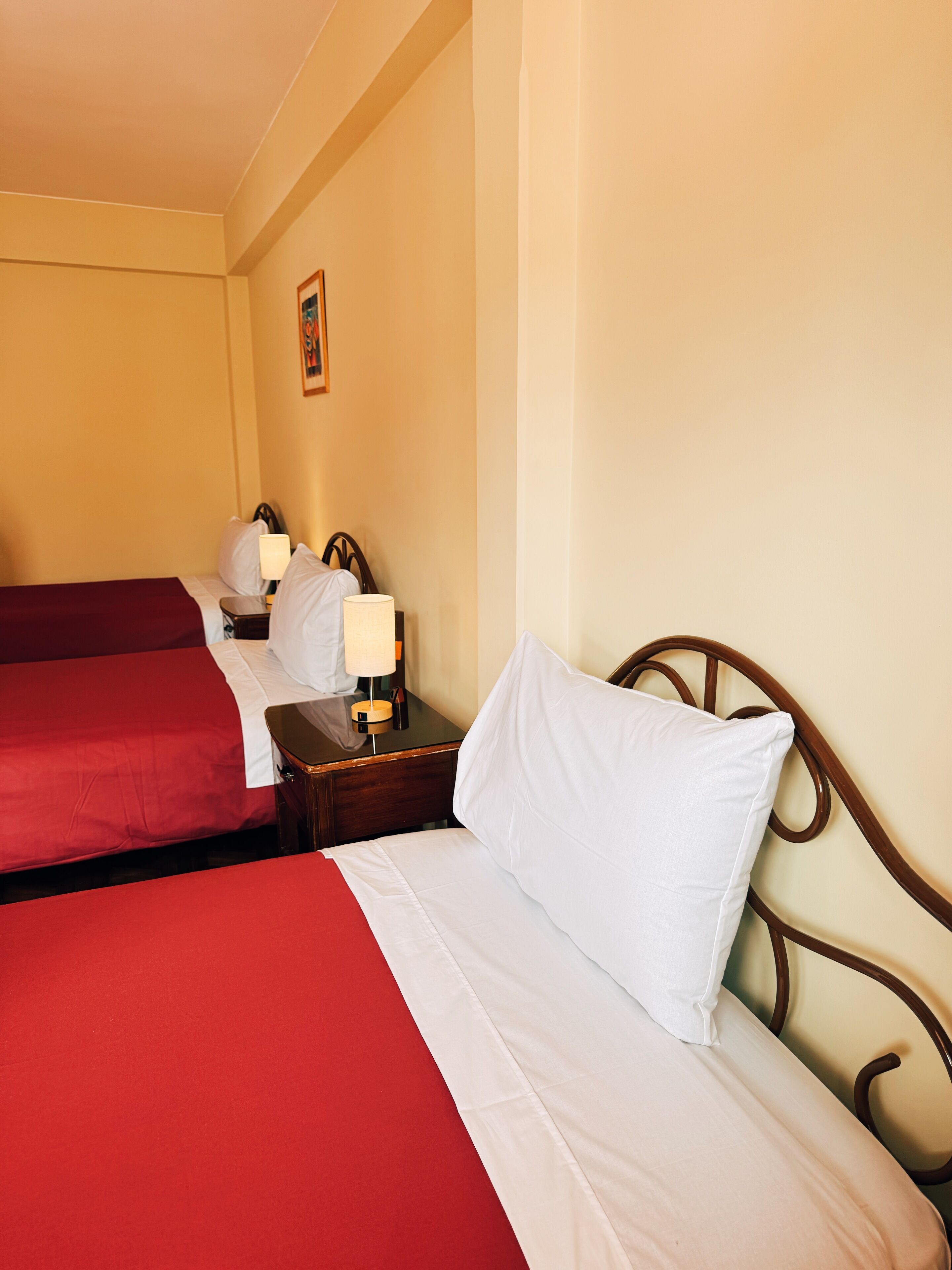 A hotel room with two beds, each with a red and white bedspread, a wooden nightstand with a lamp, and a framed picture on the wall.