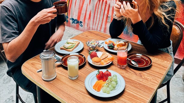 Deux personnes sont assises à une table en bois devant un repas composé de pain grillé, de fruits et de boissons, et en arrière-plan, on peut voir une fresque murale aux couleurs vives.