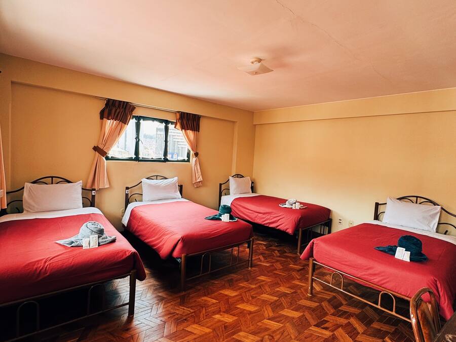 Quarto com quatro camas, cada uma com roupa de cama vermelha, piso de madeira e uma janela com cortinas.