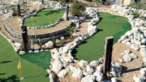 A golf course with a putting green, surrounded by rocks and palm trees, with a building in the background.