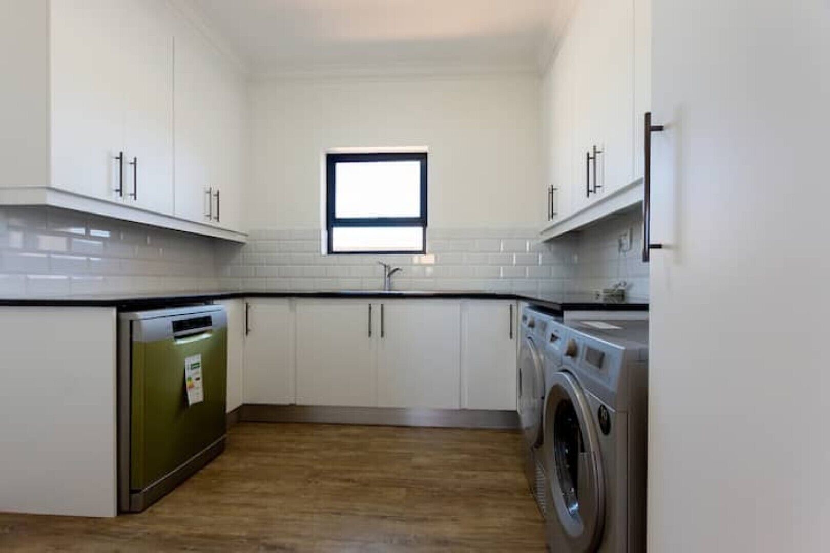 A compact kitchen with white cabinets, a built-in dishwasher, a washing machine, and a window above the sink.