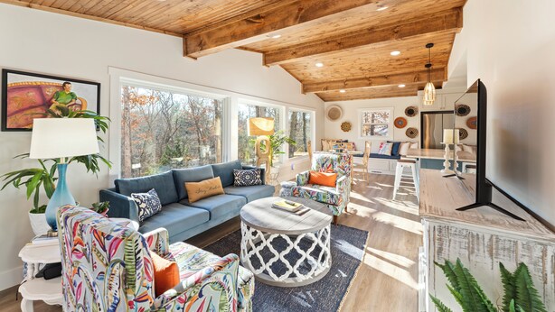 Ein großzügiges Wohnzimmer mit Holzboden, großen Fenstern, einem blauen Sofa, bunten Stühlen und einem runden Couchtisch.