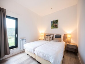 Een tweepersoonsbed met witte beddengoed, twee nachtkastjes met lampen, een raam met gordijnen, een radiator en een aan de muur hangend schilderij.