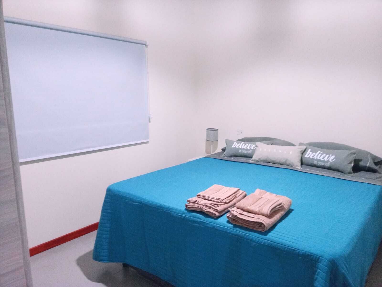 Una habitación con una cama cubierta por una colcha azul, dos almohadas con la palabra "believe" y una pila de toallas dobladas.