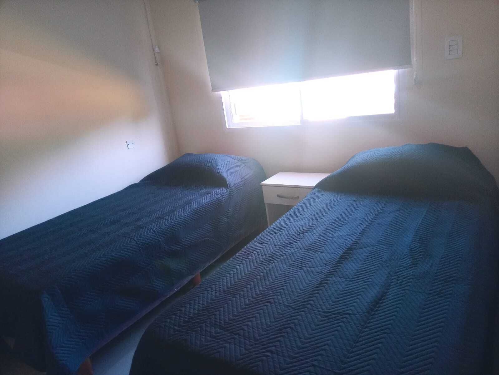 Une petite pièce avec deux lits simples, un bureau blanc et une fenêtre avec des stores.