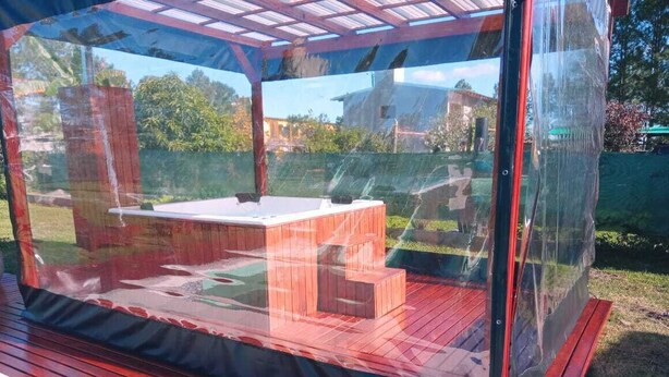 Un espacio de estar al aire libre cubierto con una mesa y bancos, rodeado por un cerramiento transparente.