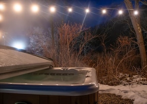 Ein Whirlpool mit Holzverkleidung, umgeben von Schnee und Bäumen, beleuchtet durch Lichterketten.