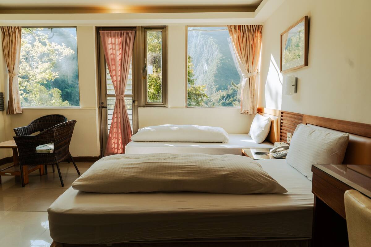A hotel room with a bed, a chair, a desk, and large windows with curtains.